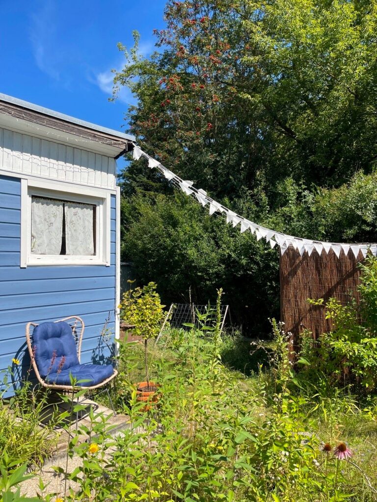 Gemütliche Sitzecke im Kleingarten mit Rattansessel und Wimpelkette vor blauem Gartenhaus