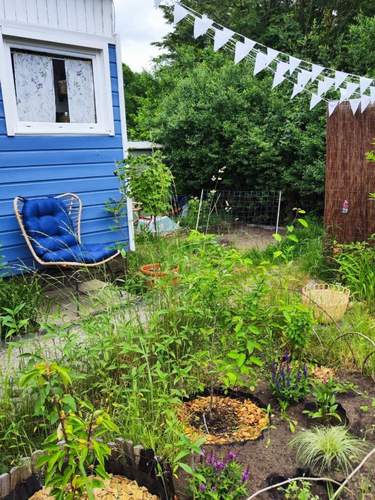 Kleingarten mit Sitzecke, Wimpelkette und naturnah bepflanzten Beeten vor blauem Gartenhaus