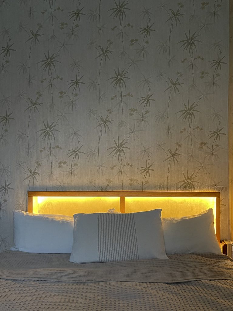 Bett mit beleuchtetem Wandlampe-DIY-Kopfteil frontal vor gemusterter Tapete